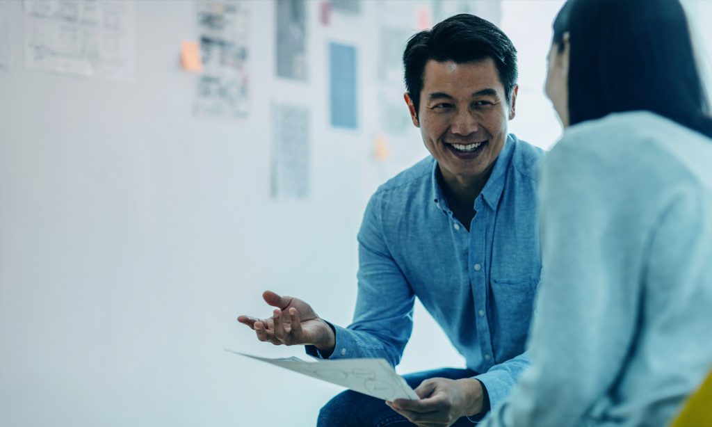 A smiling man gestures while holding a paper, engaged in conversation with a woman whose back is facing the camera.