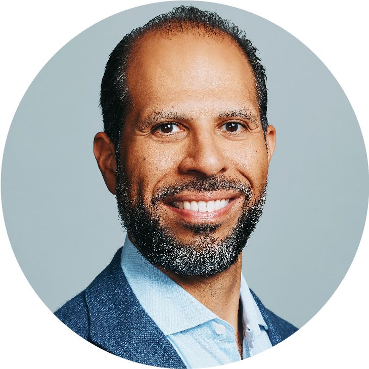 Smiling man with short hair and a trimmed beard, wearing a light blue shirt and blue blazer, posed against a plain light gray background in a circular frame, representing radiology partners subspecialty leadership.