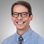 A man with short brown hair and glasses smiles at the camera. He is wearing a blue and white striped shirt and a plaid tie, against a plain gray background, giving the impression of a professional in radiology.