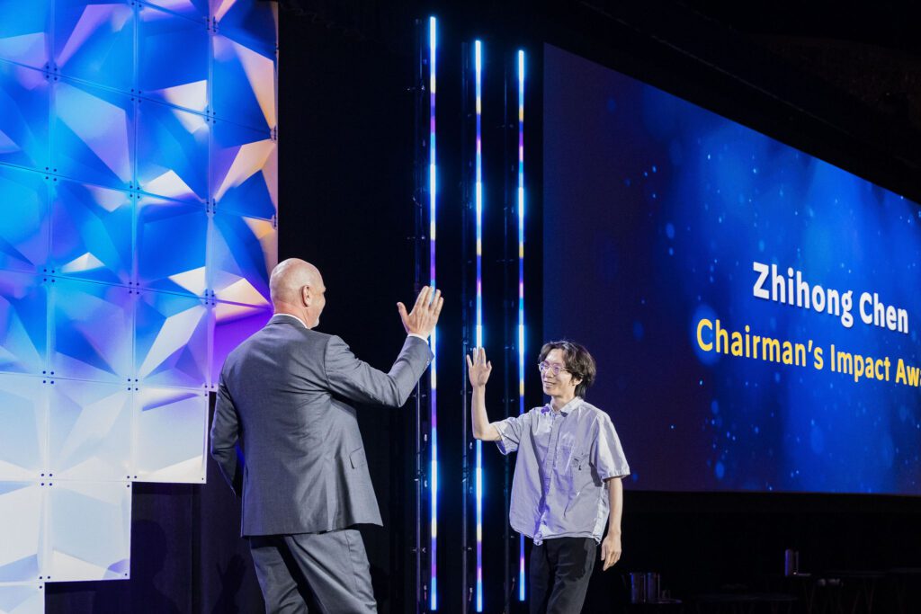 A man in a suit and a man in a short-sleeved shirt high-five on stage at an awards event. A large screen behind them displays Zhihong Chen and Chairmans Impact Award.