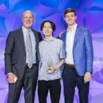 Three men stand together smiling in front of a geometric purple backdrop. The man in the middle holds a glass award. All are dressed in business attire, with jackets and collared shirts.
